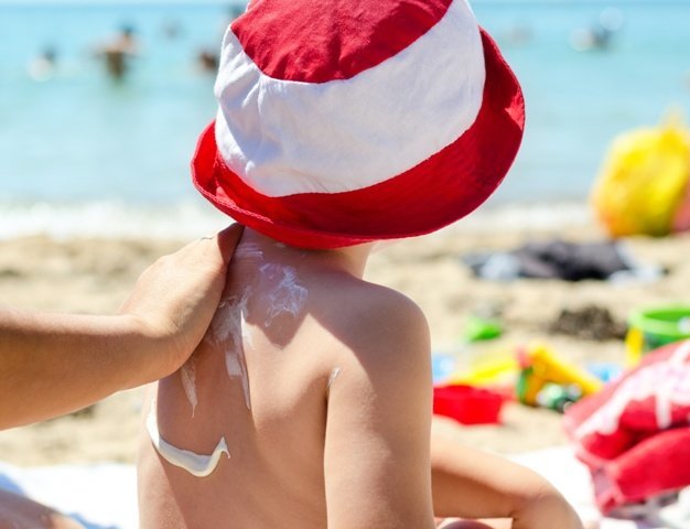 15872045 - young boy sitting on the beach during his summer vacation having sunscreen applied to his back by a parent to prevent burning from harmful uv rays