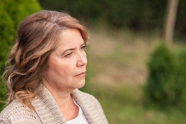 Pretty old woman is standing and resting in park. She is dreaming and looking aside pensively. Copy space in right side