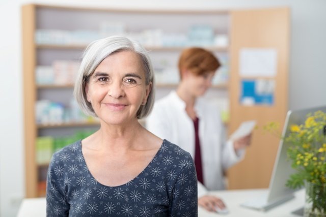 Smiling elderly patient in a pharmacy, head and shoulder facing the camera with a young female pharmacist working in the background
